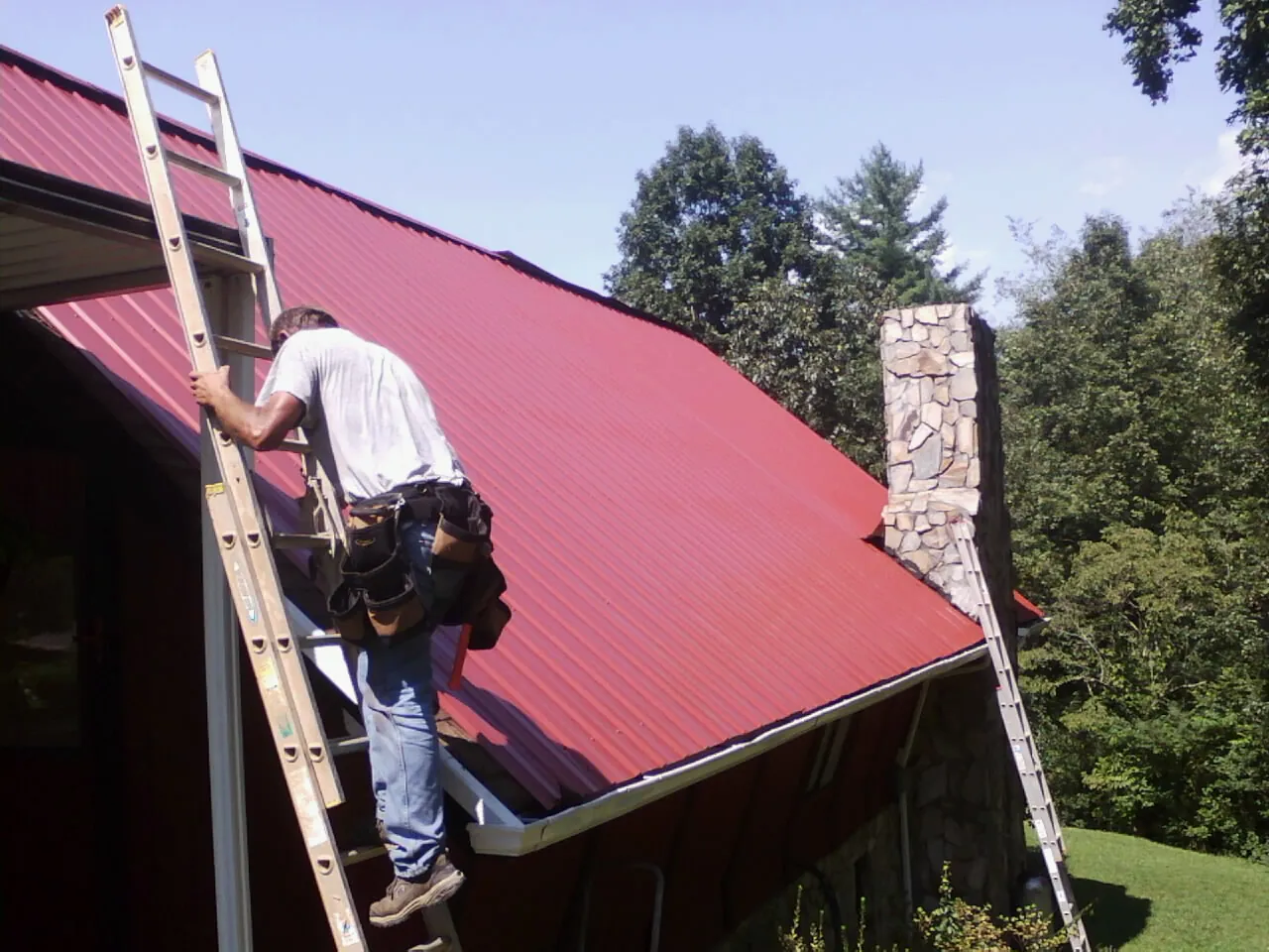 Roofer on a ladder beside a red metal roof and stone chimney — Soffit & Fascia Repair by Roofing Brothers in Fallsburg